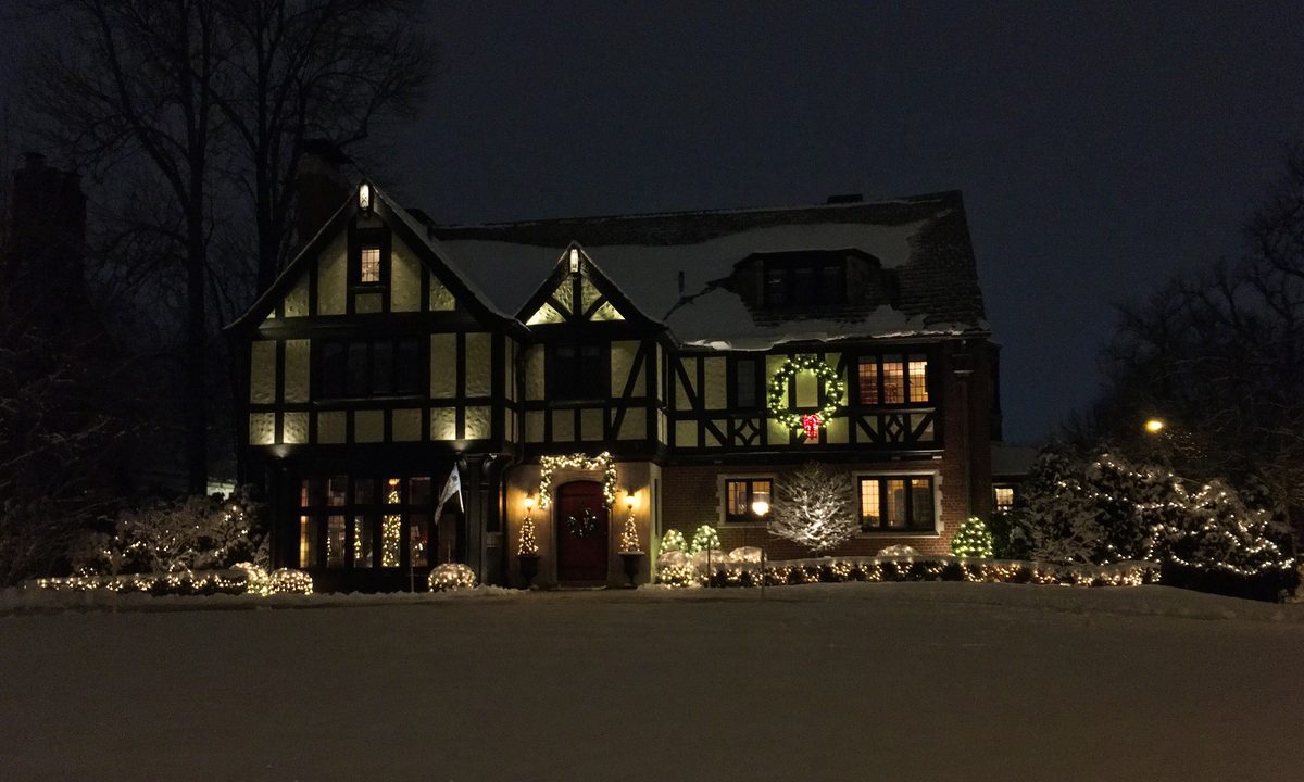 A Tudor-style house at night with Christmas lights on the shrubs, a lit wreath beside the upstairs windows and snow on the lawn and roof